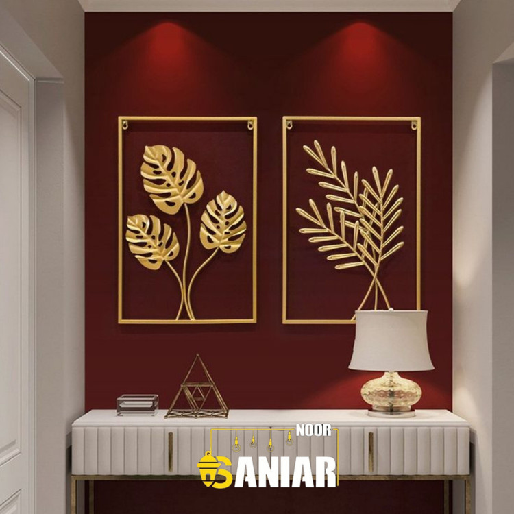 نمای نزدیک از ست دو تایی تابلو دیواری طلایی طرح برگ استوایی نصب شده روی دیوار قرمز بالای کنسول سفید».