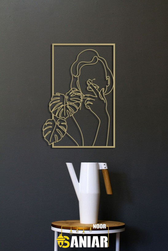 تابلو دکوراتیو لاینآرت طلایی با طرح چهره زن و برگهای انجیری نصب شده روی دیوار تیره رنگ بالای یک گلدان مدرن سفید».