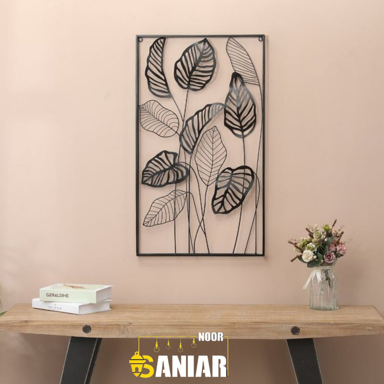 تابلو دکوراتیو مشکی با طرح برگهای پهن گیاهی نصب شده روی دیوار کرم رنگ در بالای یک میز چوبی مدرن به همراه گلدان گل».