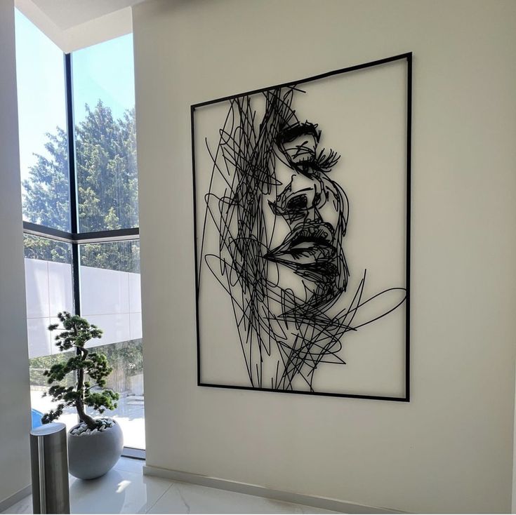 تابلو فلزی بزرگ مشکی با طرح چهره زن به سبک خطوط درهم‌پیچیده (Scribble) نصب شده روی دیوار سفید در یک فضای مدرن با پنجره بزرگ و بونسای کنار آن».