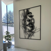 تابلو فلزی بزرگ مشکی با طرح چهره زن به سبک خطوط درهمپیچیده (Scribble) نصب شده روی دیوار سفید در یک فضای مدرن با پنجره بزرگ و بونسای کنار آن».
