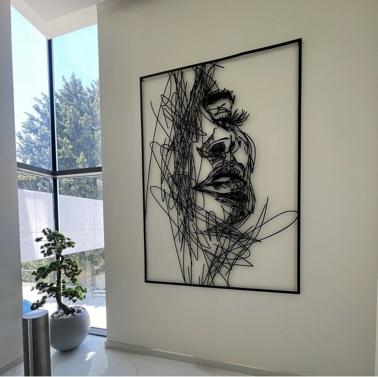 تابلو فلزی بزرگ مشکی با طرح چهره زن به سبک خطوط درهمپیچیده (Scribble) نصب شده روی دیوار سفید در یک فضای مدرن با پنجره بزرگ و بونسای کنار آن».