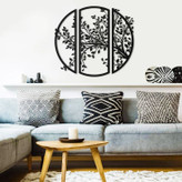 ست سه تایی دیوارکوب مشکی فلزی که در کنار هم طرح یک دایره بزرگ با شاخ و برگهای درخت را تشکیل میدهند، نصب شده روی دیوار سفید بالای کاناپه طوسی».