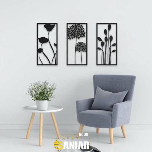 ست سه تایی قاب فلزی مشکی با طرحهای متفاوت گیاهی و شکوفه، نصب شده روی دیوار سفید بالای یک مبل تک نفره طوسی و میز سفید کوچک».