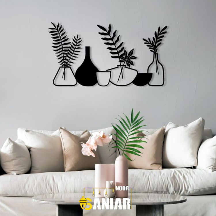 دیوارکوب فلزی مشکی با طرح چندین گلدان فانتزی و برگهای سرخس که به صورت افقی روی دیوار خاکستری روشن بالای کاناپه سفید نصب شده است».