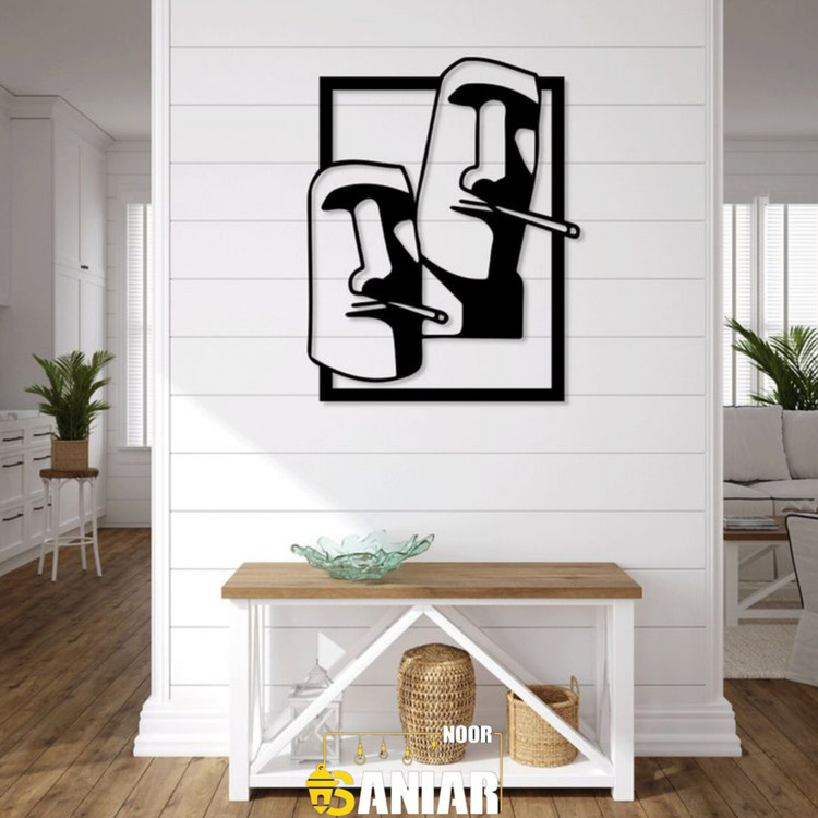 یک تابلو فلزی مشکی با طرح دو چهره سنگی موآی در یک قاب مستطیلی، نصب شده روی دیوار سفید بالای یک میز کنسول مدرن چوبی».