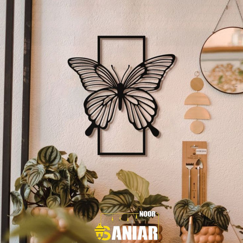 یک دیوارکوب فلزی مشکی با طرح پروانه که بالهایش از یک قاب مستطیلی بیرون زده است، نصب شده روی دیوار کرم در کنار گلدانهای گیاه پتوس و یک آینه گرد».