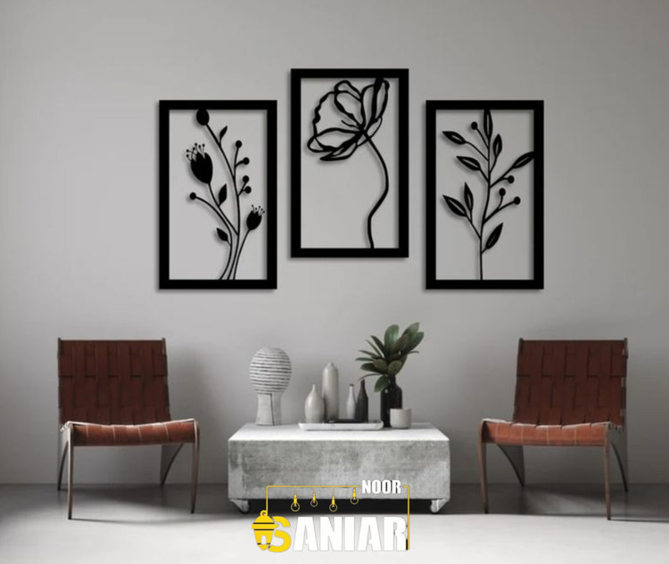 سه عدد قاب فلزی مشکی با طرحهای متفاوت ساقه و گل، نصب شده با اختلاف ارتفاع روی دیوار طوسی روشن بالای میز جلو مبلی مدرن».