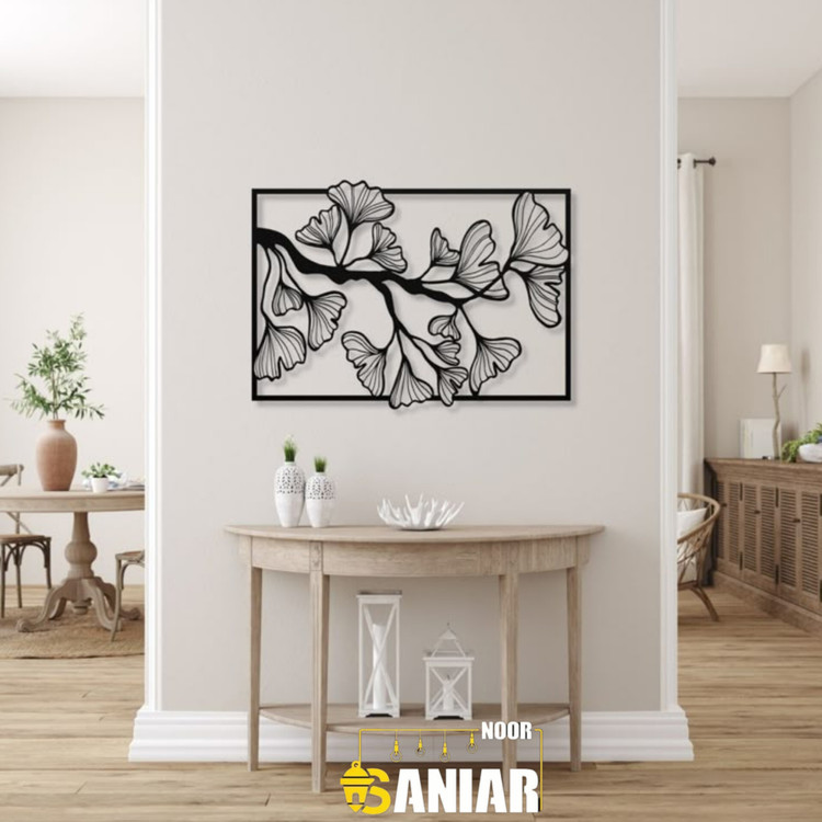 یک تابلو فلزی مشکی بزرگ با طرح شاخه و برگهای ژینکو، نصب شده روی دیوار کرم روشن در فضای بالای یک میز کنسول چوبی مدرن».