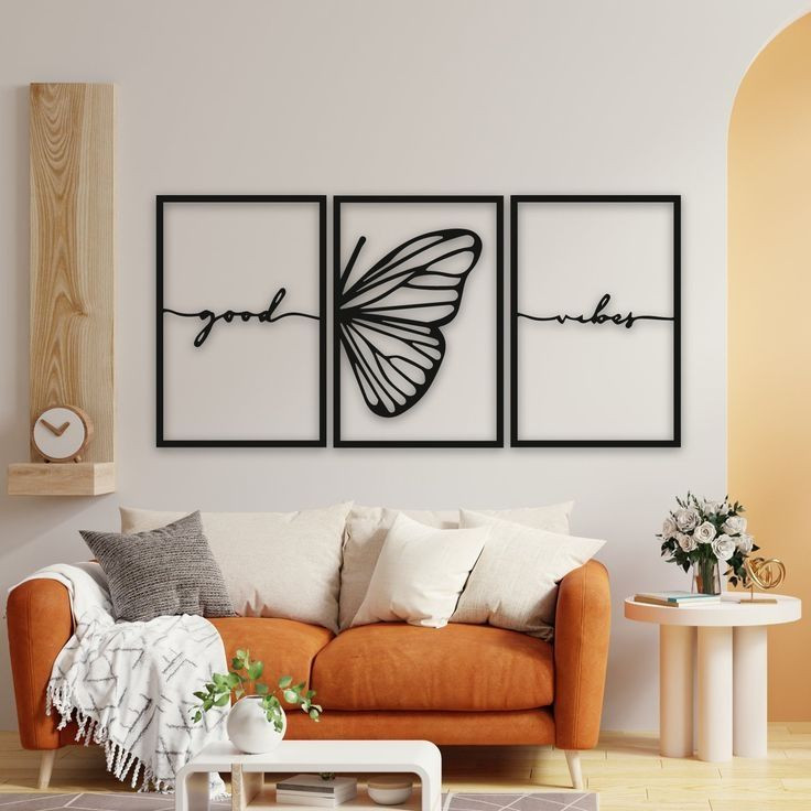 ست سه تایی قاب فلزی مشکی شامل کلمات Good و Vibes که به بالهای یک پروانه در قاب وسط متصل شدهاند، نصب شده روی دیوار سفید بالای یک کاناپه نارنجی مدرن».