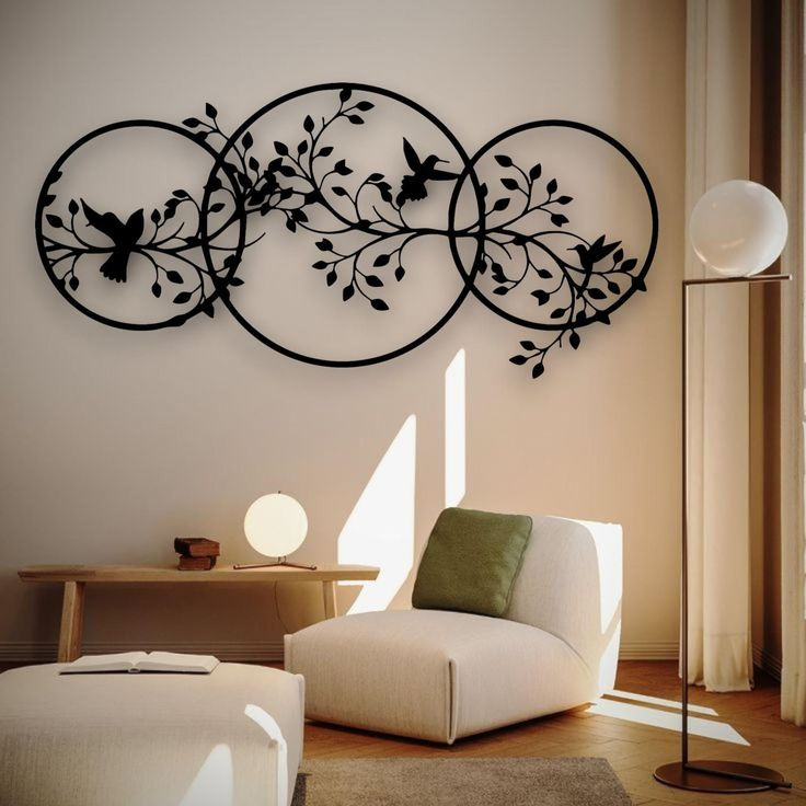 یک تابلو فلزی مشکی بزرگ شامل سه دایره متصل به هم که شاخههای درخت و پرندگان از میان آنها عبور کردهاند و روی دیوار کرمرنگ یک اتاق نشیمن مدرن نصب شده است.»